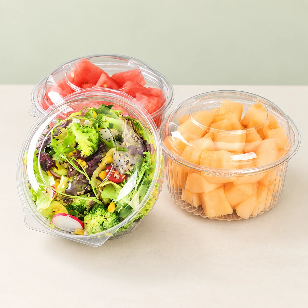 Transparent bowls containing fresh salad, watermelon and cantaloupe on a light background